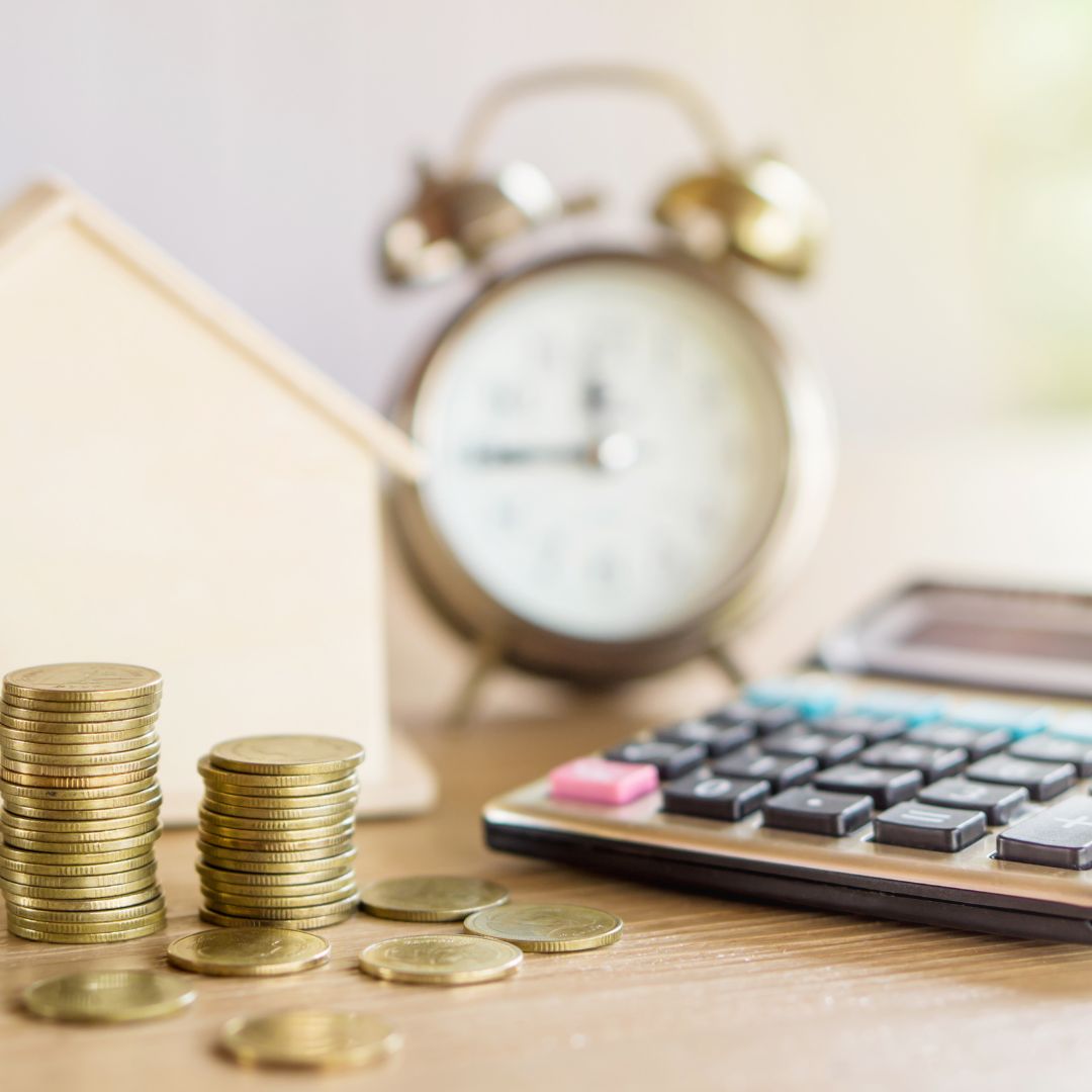 Stack of coins next to a wooden model house, clock, and a calculator. 
